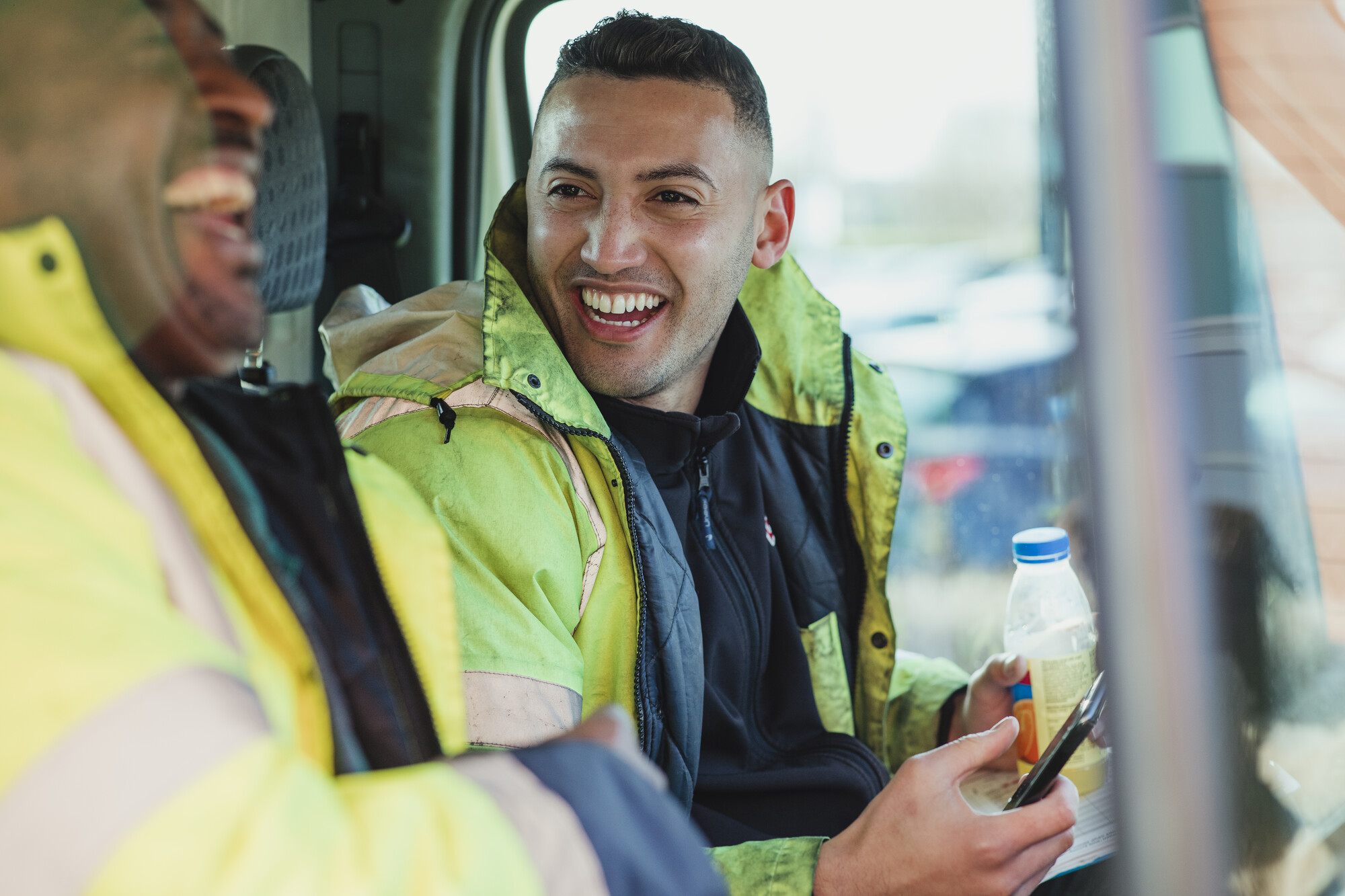 Two Male Labourers Wearing High Vis Jackets Talking And Laughing In Their Work Van Together