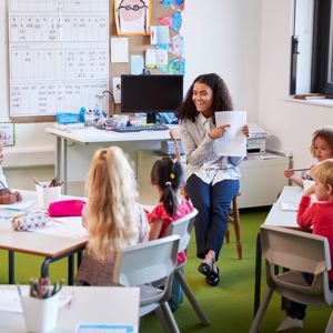 Female primary school teacher sitting on a chair facing school children in a classroom holding up and explaining a worksheet to them