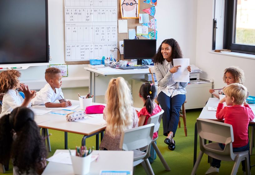 Female primary school teacher sitting on a chair facing school children in a classroom holding up and explaining a worksheet to them