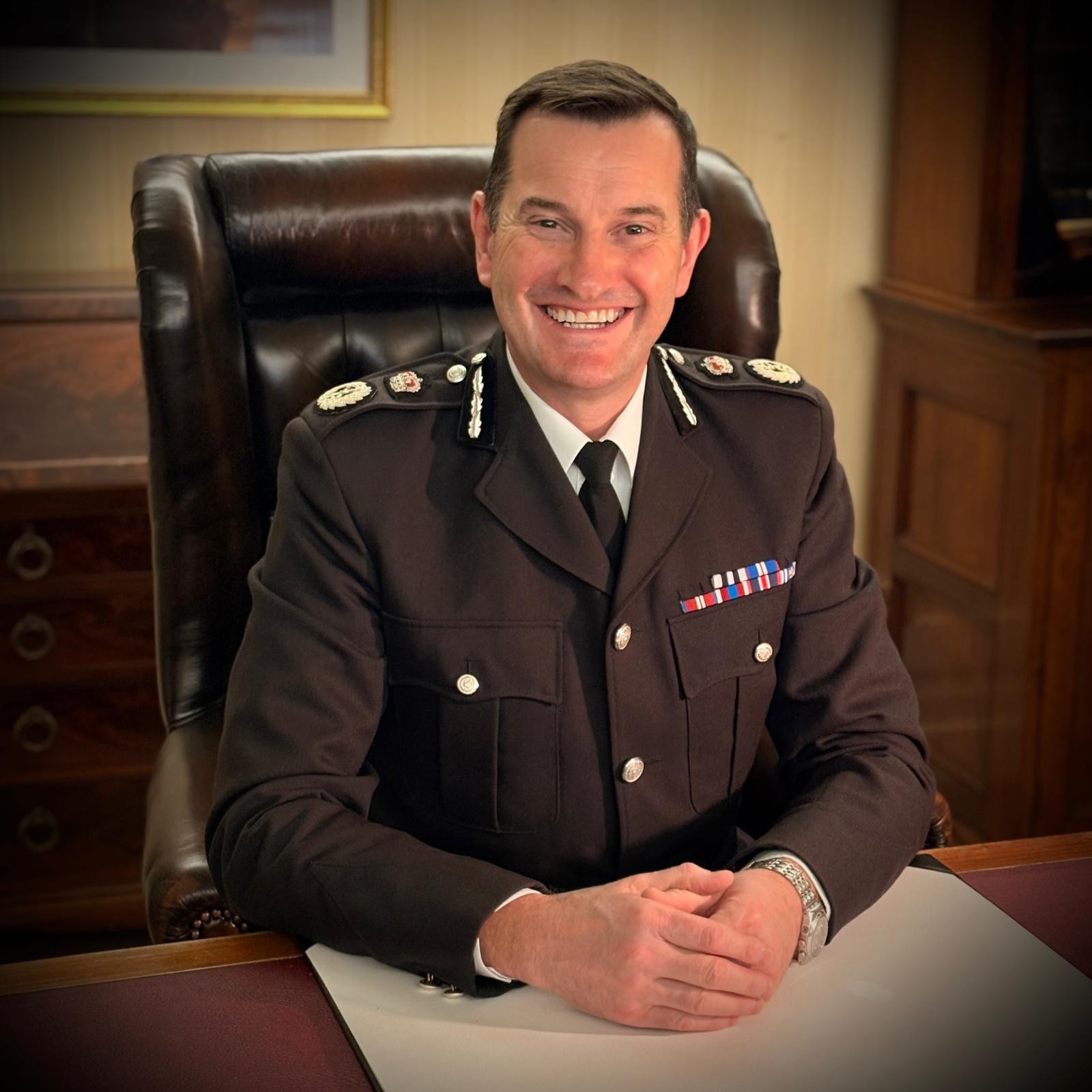 Sir John Robins in uniform smiling sitting at desk in a brown chair with his hands on the table in front of him